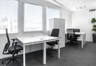 Bright shared office with white desks, ergonomic chairs, and large windows with blinds.