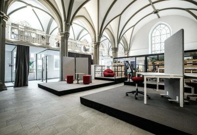 Spacious coworking lounge with raised platforms, grey carpeting, and white desks under high arched ceilings.