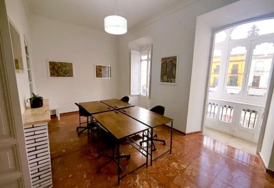 Bright coworking area with clustered desks, large window, and warm terracotta floor tiles.