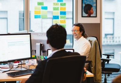 Bright open-plan office with employees at desks and a wall of colorful sticky notes.