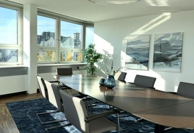 Sunlit boardroom with a large oval table, blue area rug, and city views through wide windows.