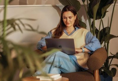 A woman working on a laptop in a cozy brown armchair surrounded by indoor plants.
