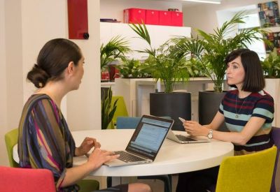 Bright communal coworking area with people working at a round table near lush green plants.