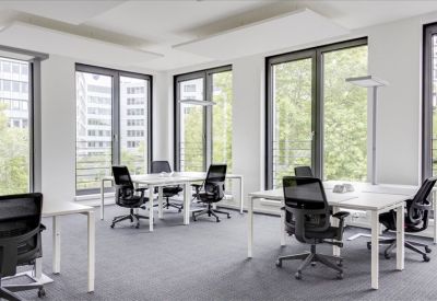 Bright open-plan office with white desks, black ergonomic chairs, and large windows overlooking trees.