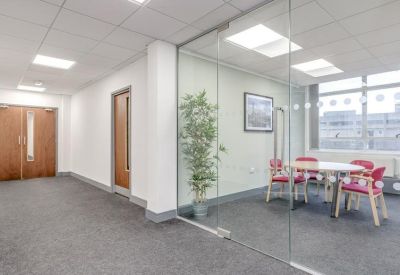 Bright hallway leading to a glass-walled meeting room with pink chairs and a potted plant.