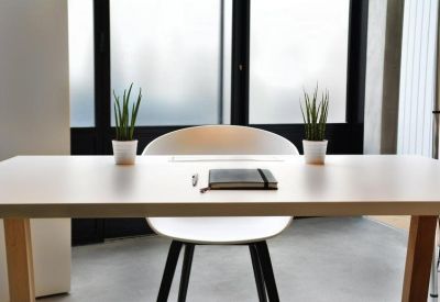 Minimalist private office desk with a white chair, notebook, and two small plants against a window.