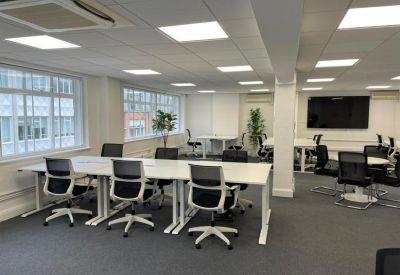 Bright workspace featuring long white communal tables and ergonomic black chairs near large windows.