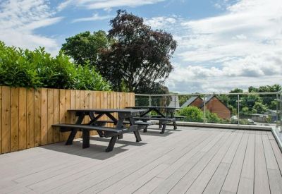 Outdoor wooden roof terrace with picnic tables and glass railings overlooking trees.