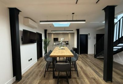 Communal breakout area with a long wooden table, grey bar stools, and a kitchen in the background.