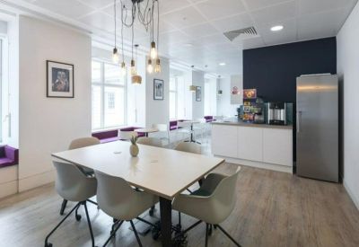 Bright breakout space with a dining table, grey chairs, and a stainless steel refrigerator.