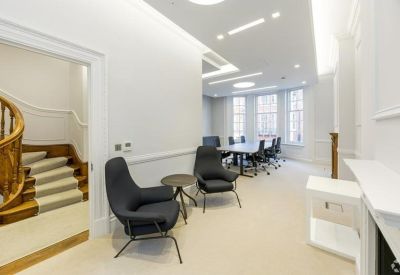 Bright breakout lounge with grey armchairs, light carpet, and a view of a spiral staircase.