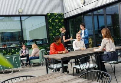 Outdoor terrace with wooden communal tables, greenery, and people socializing in the sun.