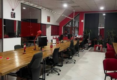 Long wooden communal desk with black office chairs and red accents under bright ceiling lights.
