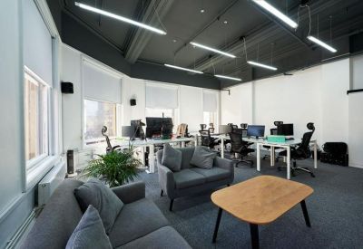 Open-plan office with grey sofas, a wooden coffee table, and workstations under a dark ceiling.