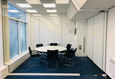 A glass-walled meeting room featuring a round white table and black chairs on blue carpet.
