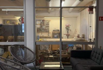 Casual lounge area with a grey sofa, woven chairs, and a glass-paneled workshop view.