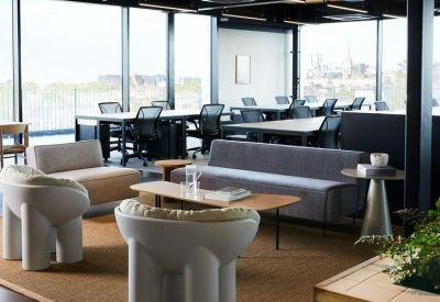 Open-plan office space with long workstations, ergonomic chairs, and a lounge area by large windows.