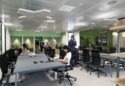 Open-plan coworking area with long grey desks, black ergonomic chairs, and modern lighting.