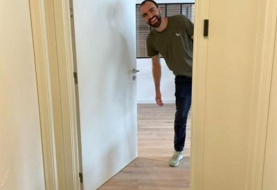 Man smiling and peeking through an open white door with a Welcome mat.