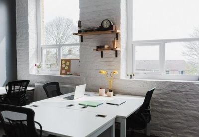 Shared office space with multiple white desks, ergonomic chairs, and large windows.