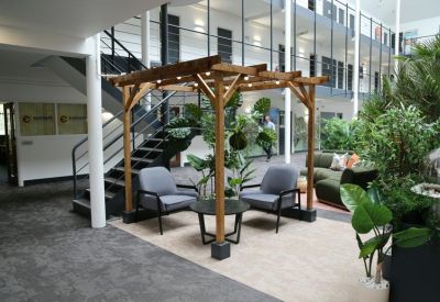Bright atrium lounge featuring a wooden pergola, grey armchairs, and lush green plants.