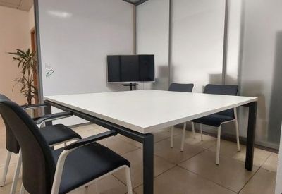 Small meeting room with a white square table, black chairs, and a wall-mounted monitor.