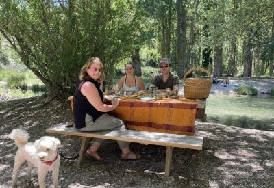 A group enjoying a picnic by a clear river under shaded trees.