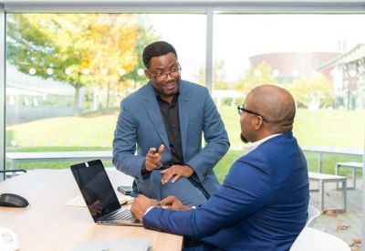 Two professionals in suits discussing work at a desk with a laptop.