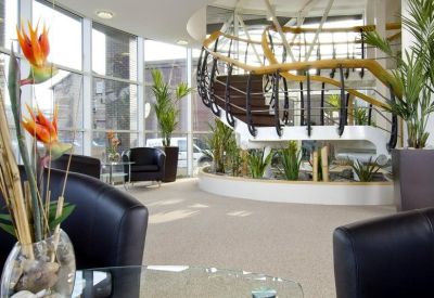 Sunlit lounge area with black leather armchairs, a glass coffee table, and an ornamental staircase.