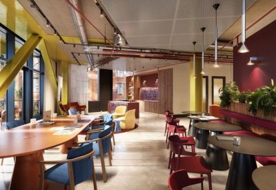 Open-plan coworking space with long wooden tables, blue velvet chairs, and a cafe area in the background.
