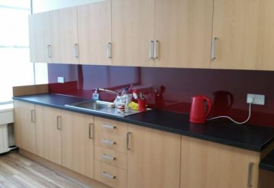 Modern communal kitchen with light wood cabinetry, dark countertops, and a deep red backsplash.