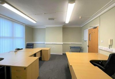 Large shared office suite with multiple wooden L-shaped desks and natural light.