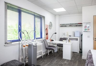 Clinical treatment room with medical equipment, desk, and grey seating by a window.