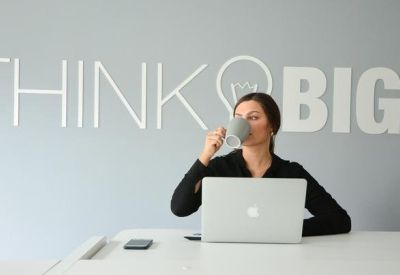 Woman drinking coffee at a desk with Think Big wall lettering.