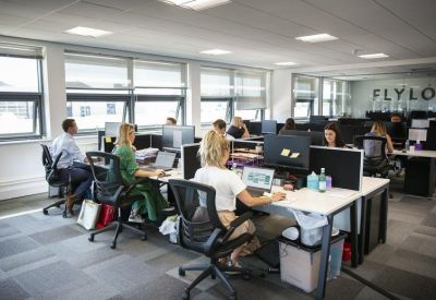 Open-plan office space with professionals working at rows of white desks by large windows.