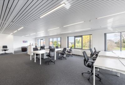 Spacious open-plan office with white desks and large windows overlooking greenery.