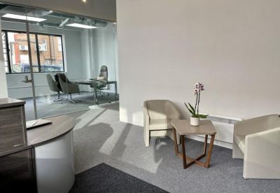 Reception lounge area with comfortable armchairs, a side table with flowers, and a view into a private office.