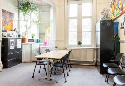 Bright communal dining area with a large wooden table, potted plants, and tall windows.