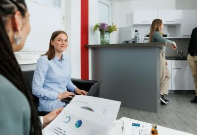 Spacious shared office area with people working at desks and a small kitchen corner in the background.
