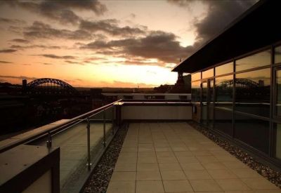 Paved outdoor terrace with glass railings overlooking a city sunset.
