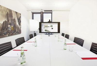 Bright conference room with a long white table, black chairs, and video conferencing screen.