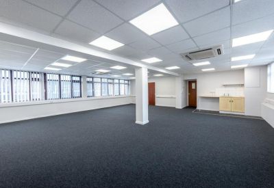 Large empty open-plan office space with grey carpeting, white pillars, and ample natural light from windows.
