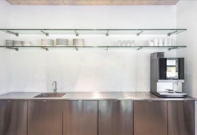 Minimalist communal kitchen area with stainless steel cabinetry and glass shelving.