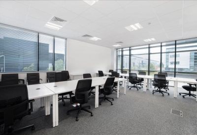Spacious open-plan workspace with rows of white desks and black ergonomic chairs under bright panel lighting.