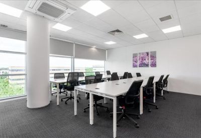 Spacious coworking area with long white tables and views of green trees through large windows.