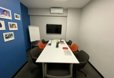 Meeting room with a long white table, various colored chairs, a wall-mounted TV, and a blue accent wall.