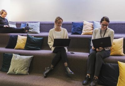 Three people working on laptops while sitting on tiered upholstered seating with colorful pillows.