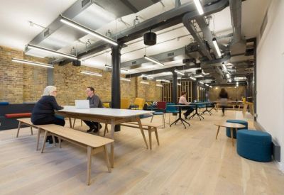 Open-plan coworking space with long wooden tables, benches, and exposed brick walls.