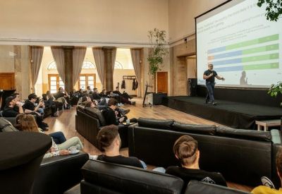 Large event hall with a presentation screen and attendees seated on sofas.