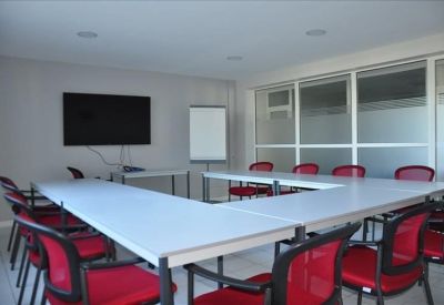 U-shaped conference table surrounded by red chairs in a bright meeting room with a wall-mounted TV.